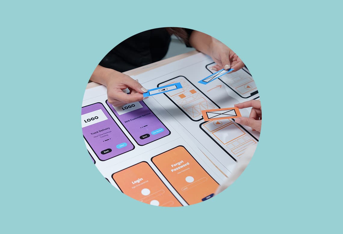 Hands collaborating on mobile app website redesign with colorful interface mockups on a table, conveying teamwork and creativity in a tech setting